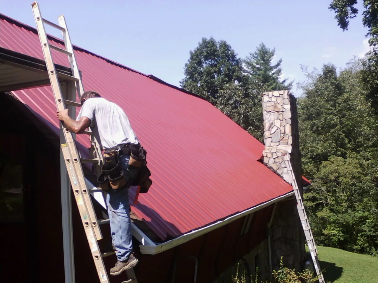 Roofer on a ladder beside a red metal roof and stone chimney — Soffit & Fascia Repair by Roofing Brothers in Big Rock